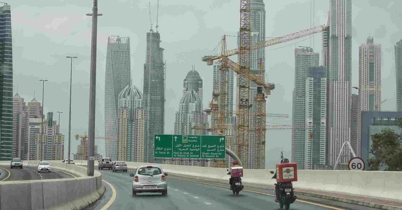 Saudi Construction Industry Outlook: Highway view with cars and delivery motorcycles, flanked by tall buildings and construction cranes under an overcast sky.
