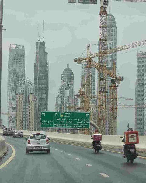 Saudi Construction Industry Outlook: Highway view with cars and delivery motorcycles, flanked by tall buildings and construction cranes under an overcast sky.
