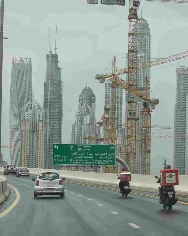 Saudi Construction Industry Outlook: Highway view with cars and delivery motorcycles, flanked by tall buildings and construction cranes under an overcast sky.