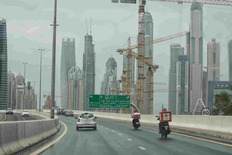 Saudi Construction Industry Outlook: Highway view with cars and delivery motorcycles, flanked by tall buildings and construction cranes under an overcast sky.